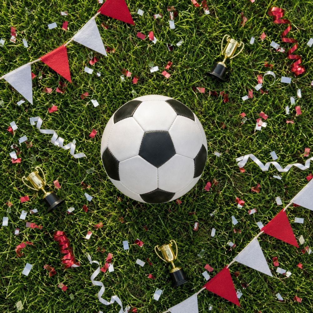 Overhead-Flatlay mit Fußball auf Naturrasen, Konfetti, Pokalen und Wimpeln in strahlendem Tageslicht